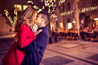 Ein verliebtes Paar küsst sich und er hebt sie leicht hoch und hat seine Arme um sie gelegt - sie wirken sehr glücklich und im Hintergrund sind Stühle von einem Cafe zu sehen und Lichterketten hüllen den Fußweg in ein orangenes und romantisches Licht 