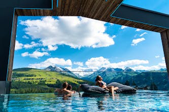 Überdachter Outdoor Swimmingpool mit 3 entspannten Personen darin - eine sitzt auf einem schwimmenden Kissen und genießt die Sonne - die anderen beiden Personen stehen zusammen im Wasser und sehen die großen gut sichtbaren Berge und Wälder - der himmel strahlt blau mit wenigen Wolken
