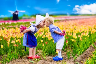 Zwei süße Kinder, die sich mit leicht gebeugten Rücken und mit Blumen in den Händen mit der Nasenspitze berühren - im Hintergrund ist eine Windmühle und die 2 Kinder stehen auf einem großen Feld voller Blumen in Holland - den Niederlanden